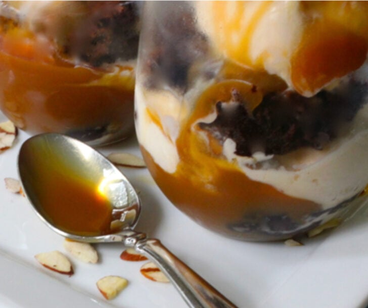 Close-up of a layered caramel dessert in a glass, with cream and almond pieces, a spoon resting on a plate nearby.