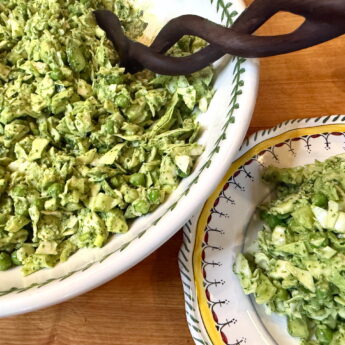 Large white bowl of chopped avocado salad with herbs, a wooden spoon resting on top, on a wooden table.