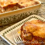Slice of cheesy baked lasagna on a decorative plate with a fork nearby, from a casserole dish in the background.