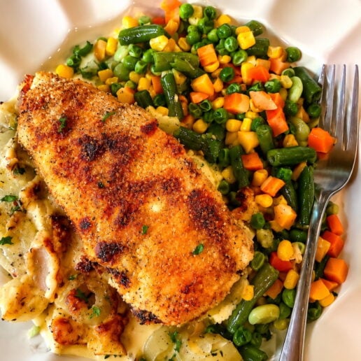 Crispy breaded fish fillet on a creamy bed with a side of mixed vegetables (peas, corn, carrots, green beans) on a white plate with a fork nearby.
