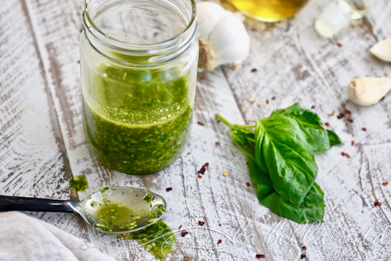 Jar of green pesto on a rustic wooden table with fresh basil, garlic, and olive oil nearby.