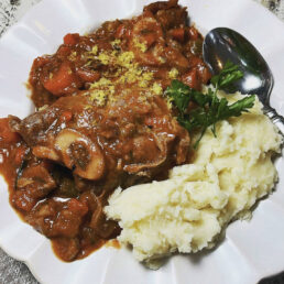 Beef stew with chunks of meat and vegetables in brown gravy, served with mashed potatoes and parsley on a white plate.
