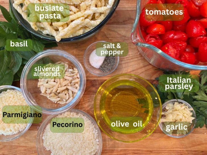 pesto alla trapanese ingredients | La Bella Vita Cucina Assorted cooking ingredients on a wooden table: basil, tomatoes, garlic, olive oil, cheeses (Parmesan and Pecorino), slivered almonds, pasta, and seasonings like salt and pepper.